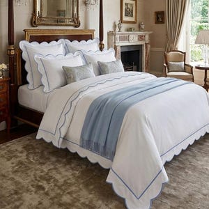 May include: A white bedding set featuring scalloped edges with light blue trim, including a duvet cover, two large pillows with matching scalloped borders, two smaller rectangular pillows with a grey pattern, and a solid light blue throw blanket.