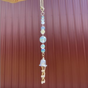 May include: A silver chain with a decorative hanging ornament. The ornament features iridescent blue and clear beads of various sizes, a bell-shaped charm, and gold-colored wire accents. The background is a reddish-brown corrugated metal surface.