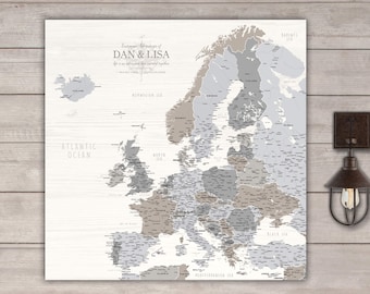 May include: A framed map of Europe with push pins, in shades of gray and brown, mounted on a white wooden background. The map is labeled with country names and ocean names. Text at the top reads "Everyone's Adventures of Dan & Lisa".