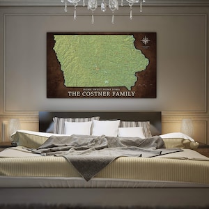 May include: A brown and green topographic map of Iowa with the text "Home Sweet Home Iowa" and "The Costner Family" printed below the map. The map is hanging on a wall above a bed with a gray and white striped bedspread.