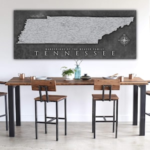 May include: A black and white map of Tennessee with the text "Wanderings of the Weaver Family" and "Tennessee" printed below the map. The map is hanging on a white wall above a wooden table with four chairs.