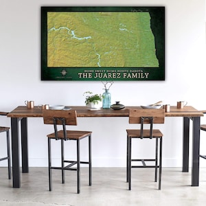 May include: A green and yellow map of North Dakota with the text "Home Sweet Home North Dakota" and "The Juarez Family" printed below the map. The map is hanging on a white wall above a wooden table with two black metal chairs.
