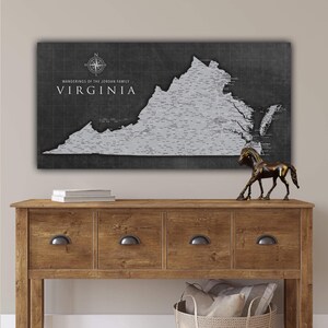 May include: A framed map of Virginia with a gray state outline and pin locations on a dark gray background. The text "Wanderings of the Jordan Family" and "Virginia" are displayed in white. The map hangs above a wooden console table.