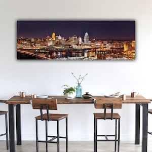 May include: A panoramic view of a city skyline at night. The city is lit up with lights, and the buildings are reflected in the water. A bridge crosses the river in the foreground.
