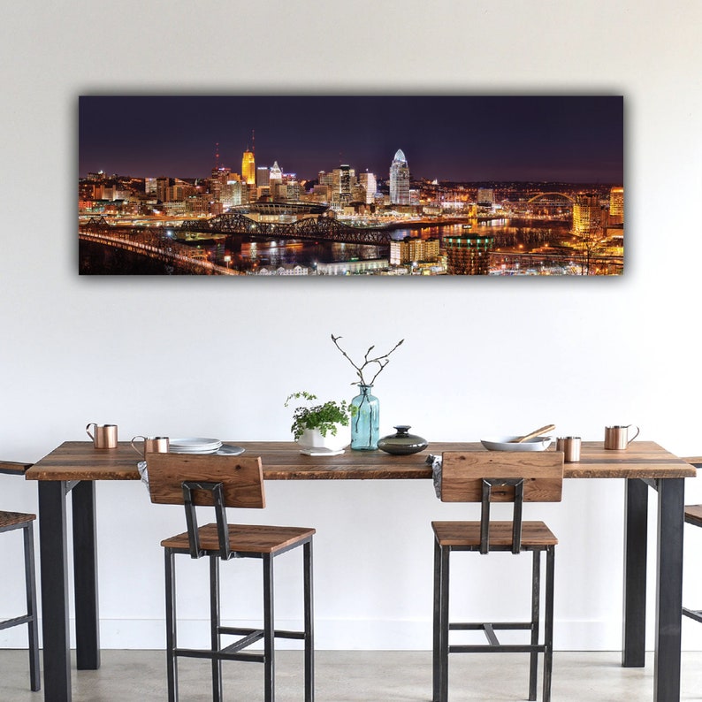 May include: A panoramic view of a city skyline at night. The city is lit up with lights, and the buildings are reflected in the water. A bridge crosses the river in the foreground.