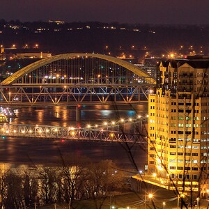 May include: A nighttime cityscape with a large building illuminated with lights and a bridge crossing a river. The bridge is lit with yellow lights and the building is a light yellow color.