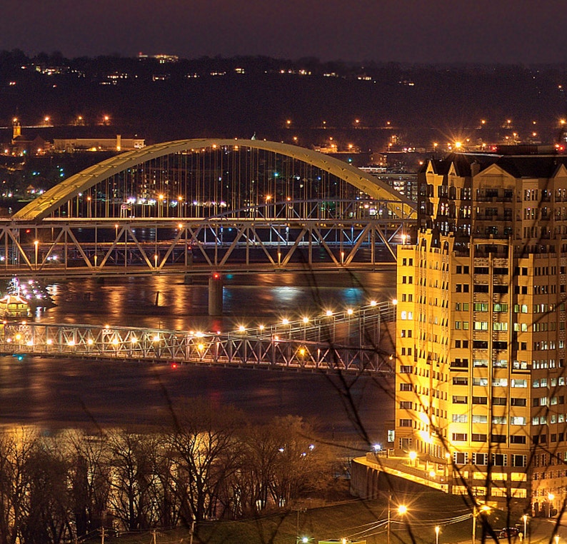 May include: A nighttime cityscape with a large building illuminated with lights and a bridge crossing a river. The bridge is lit with yellow lights and the building is a light yellow color.