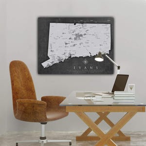 Peut inclure: Une carte de l'état du Connecticut en noir et blanc avec le texte "EVANS HOME SWEET HOME CONNECTICUT" imprimé en dessous. La carte est accrochée à un mur blanc au-dessus d'un bureau en verre avec une base en bois. Un fauteuil de bureau en cuir marron se trouve devant le bureau.