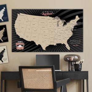 May include: A black and white map of the United States with a beige outline of the states. The map has a baseball theme with the text "Take me out to the Ball Game" and "Jason's Baseball Stadium Tour". The map is on a wall above a black desk with a laptop and a lamp.