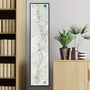 May include: A framed map print with a black border. The map is a detailed topographical representation, featuring green, white, and blue hues. The top right corner has a green circular symbol with an arrow. The bottom right corner has text that reads "Scenic Trails".