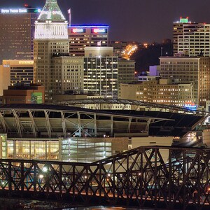 May include: A night view of a city skyline with a large stadium in the foreground. The stadium has a white roof and a metal frame. The city skyline is lit up with lights, and there are several tall buildings in the background. The buildings are all lit up with lights, and there are several tall buildings in the background.