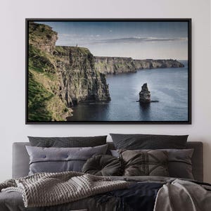May include: A framed canvas print of the Cliffs of Moher in Ireland. The image shows a dramatic coastal landscape with towering cliffs, green grass, and the deep blue ocean. A small rock formation stands in the water. The sky is a pale blue with some clouds.