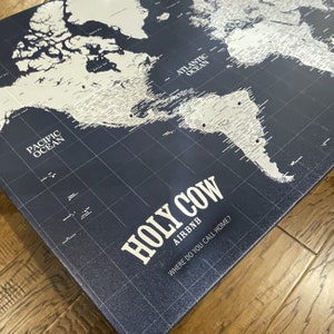 May include: A dark blue world map with white countries and cities. The map is on a canvas with a grid pattern. The text "HOLY COW AIRBNB WHERE DO YOU CALL HOME?" is printed on the bottom of the map.