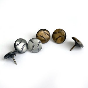 May include: Five baseball-themed tacks with a black stitching design. Two are silver, two are gold, and one is black.