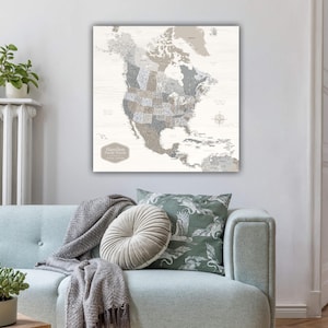 May include: A framed map of North America in neutral tones, showing state boundaries and pin locations. The map is set against a light wood-effect background. The text "Hamilton Family Travels" is visible. The map is displayed above a light blue sofa, with cushions.