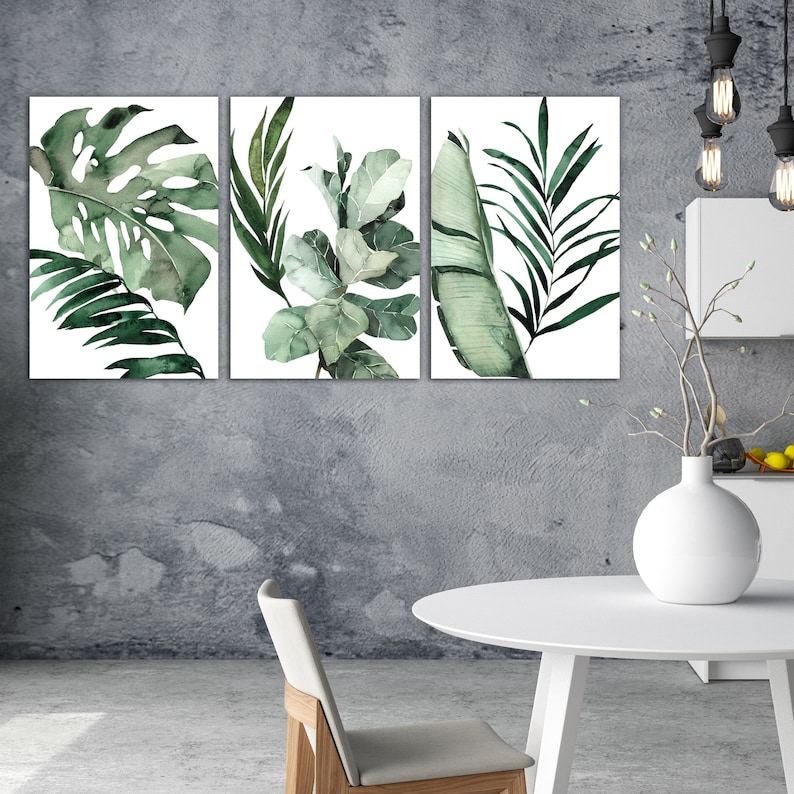 May include: Three watercolor paintings of tropical leaves in shades of green. The paintings are framed and hung on a gray wall.
