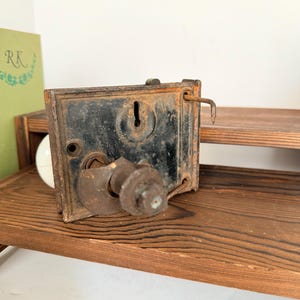 Pode incluir: Um mecanismo de fechadura de porta retangular de metal com uma placa central preta com buraco de fechadura, um conjunto de maçaneta de metal enferrujado saliente e um pequeno pino de metal dobrado que se estende do canto superior direito.