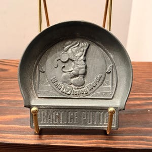 May include: A circular pewter-colored metal tray featuring a raised relief of Mickey Mouse playing golf. The text Walt Disney World is embossed below the figure, and the words PRACTICE PUTTER are engraved on a rectangular base plate.