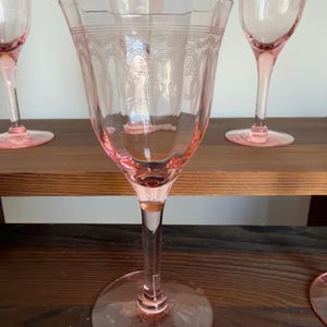 May include: A set of pink glass goblets featuring a delicate etched pattern around the upper rim. Each goblet has a slender stem and a wide circular base, with a translucent pink hue throughout the glass structure and a scalloped edge design.