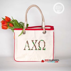 May include: A cream-colored canvas tote bag with red trim and two beige rope handles. The front features the Greek letters AXΩ in a green and brown floral pattern. A bouquet of orange tulips is tucked inside the bag, and a circular logo is visible.