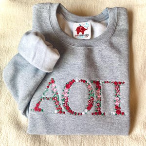 May include: A heather gray crewneck sweatshirt featuring the Greek letters AOII embroidered in a floral pattern. The letters are composed of small red, pink, and white flowers with green leaves. A white tag with a red elephant logo reads redelephant.com.