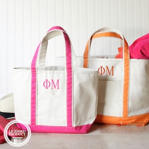 May include: Two off-white canvas tote bags with dual handles. The left bag features bright pink trim and the pink Greek letters ΦM. The right bag has orange trim and the orange Greek letters ΦM. Both bags have a front pocket and a flat base.