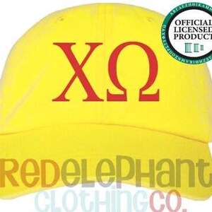 May include: A bright yellow baseball cap featuring the red Greek letters Chi and Omega centered on the front. A circular green and white seal in the upper right corner displays the text Official Licensed Product. The text RedElephant Clothing Co.