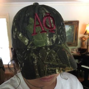 May include: A baseball cap featuring a camouflage pattern in shades of dark green, brown, and black. The front of the cap displays the Greek letters Alpha and Phi embroidered in a bold, dark red thread with a distinct stitched texture.