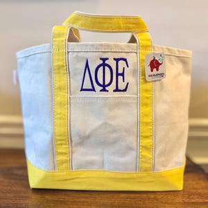 May include: A white canvas tote bag with yellow trim and handles. The front features the Greek letters Delta Phi Epsilon embroidered in purple thread. A small square tag with a red elephant logo and text is attached to the upper right side.