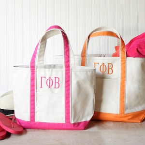 May include: Two off-white canvas tote bags with open tops and double handles. The left bag features bright pink trim and the Greek letters ΓΦΒ in pink. The right bag has orange trim and the Greek letters ΓΦΒ in orange. Both bags have a front pocket.