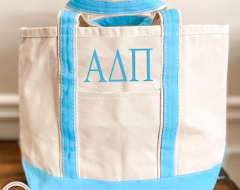 May include: A beige canvas tote bag featuring bright blue trim on the base and handles. The front of the bag displays the Greek letters Alpha Delta Pi in a matching bright blue font. The bag includes an open exterior pocket on the front side.