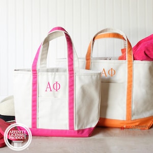 May include: Two off-white canvas tote bags with open tops and dual handles. The left bag features bright pink trim and the pink Greek letters ΑΦ. The right bag has orange trim, orange Greek letters ΑΦ, and a bright pink fabric item inside.