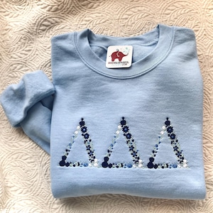 May include: A light blue crewneck sweatshirt featuring three embroidered Greek delta symbols arranged horizontally. Each triangle is composed of small dark blue, light blue, and white floral patterns. A white tag with a red elephant logo and the text "REDELEPHANT.COM" is attached at the neckline.