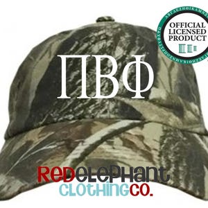 May include: A camouflage baseball cap featuring the white Greek letters Pi Beta Phi on the front. The cap includes a circular green and white seal in the upper right corner that reads Official Licensed Product and the text RedElephant Clothing Co.
