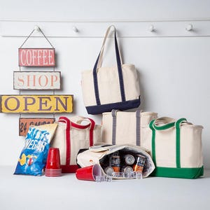Peut inclure: Une collection de sacs fourre-tout en toile cr&egrave;me avec des bordures bleu marine, rouges, grises et vertes. Un sac est ouvert, r&eacute;v&eacute;lant des canettes en aluminium et des gla&ccedil;ons &agrave; l'int&eacute;rieur. Un paquet de chips et des gobelets rouges sont pos&eacute;s.