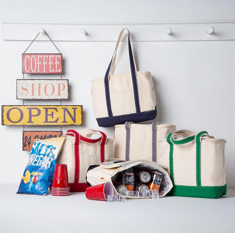 Peut inclure: Une collection de sacs fourre-tout en toile cr&egrave;me avec des bordures bleu marine, rouges, grises et vertes. Un sac est ouvert, r&eacute;v&eacute;lant des canettes en aluminium et des gla&ccedil;ons &agrave; l'int&eacute;rieur. Un paquet de chips et des gobelets rouges sont pos&eacute;s.