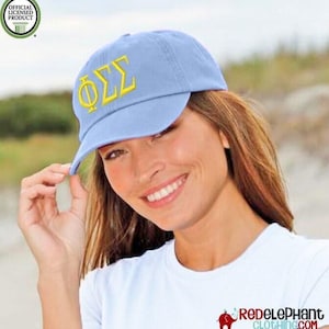 May include: A light blue baseball cap featuring the Greek letters Phi Sigma Sigma embroidered in yellow thread on the front. The cap includes an official licensed product seal in the upper left corner and a logo in the bottom right corner.