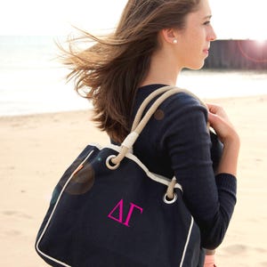 May include: A navy blue canvas tote bag featuring thick beige rope handles, silver-tone metal grommets, and white piping trim. The front of the bag displays the Greek letters Delta Gamma in pink embroidery. An Affinity Licensed Product logo is visible.