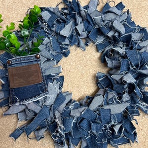 May include: A circular wreath constructed from numerous frayed strips of blue denim fabric. A single denim pocket featuring a brown leather Wrangler brand patch is attached to the left side, containing a small sprig of green artificial leaves.