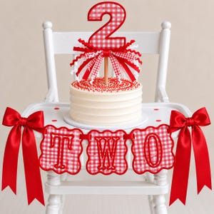 May include: A white frosted cake topped with a red and white gingham number two cake topper featuring ribbon accents. A banner with red and white gingham scalloped squares spells out the word TWO, decorated with large red satin bows on each side.