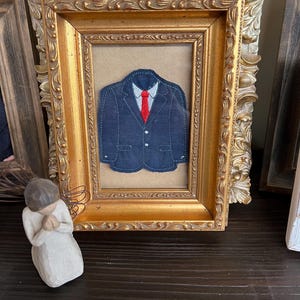 Puede incluir: Una aplicaci&oacute;n de tela de una chaqueta de traje azul oscuro con una camisa de cuello azul claro y una corbata roja brillante, exhibida dentro de un marco rectangular dorado ornamentado con detalles florales y volutas en los bordes.