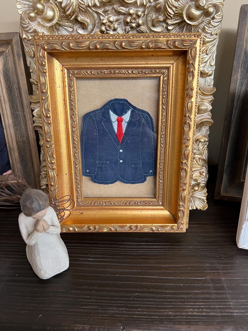 Puede incluir: Una aplicaci&oacute;n de tela de una chaqueta de traje azul oscuro con una camisa de cuello azul claro y una corbata roja brillante, exhibida dentro de un marco rectangular dorado ornamentado con detalles florales y volutas en los bordes.