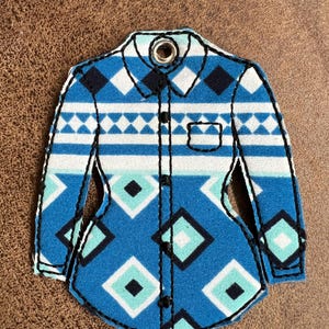Puede incluir: Un adorno de fieltro con forma de camisa que presenta un patr&oacute;n geom&eacute;trico de diamantes en azul, blanco y turquesa. El dise&ntilde;o incluye un cuello con un ojal met&aacute;lico, una tapeta frontal con costuras negras y un bolsillo en el pecho.
