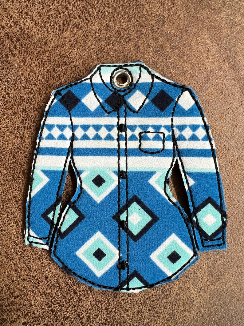 Puede incluir: Un adorno de fieltro con forma de camisa que presenta un patr&oacute;n geom&eacute;trico de diamantes en azul, blanco y turquesa. El dise&ntilde;o incluye un cuello con un ojal met&aacute;lico, una tapeta frontal con costuras negras y un bolsillo en el pecho.