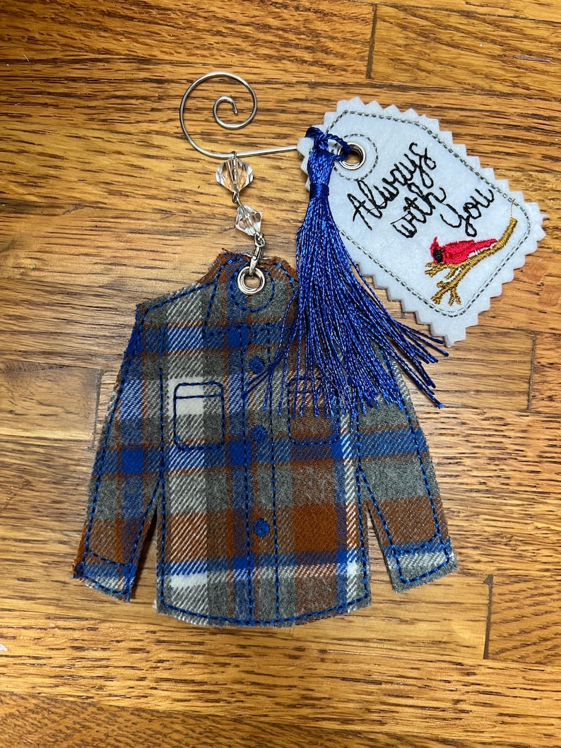 Puede incluir: Un adorno de tela con forma de camisa a cuadros en azul, marr&oacute;n y gris, con un colgador de metal plateado y cuentas transparentes. Incluye una etiqueta blanca festoneada con el texto Always with You y un cardenal rojo, adem&aacute;s de una borla azul.