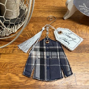 Puede incluir: Un adorno de tela con forma de camisa de manga larga con botones y un patr&oacute;n de cuadros en negro, gris y blanco. Cuenta con un colgador de metal plateado, una borla gris claro, cuentas transparentes y una etiqueta de fieltro festoneada blanca con el texto Love Always.