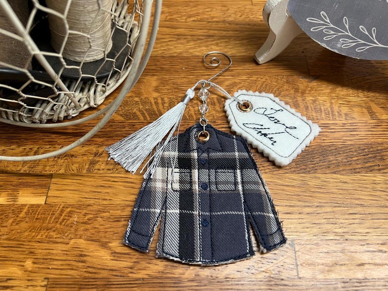 Puede incluir: Un adorno de tela con forma de camisa de manga larga con botones y un patr&oacute;n de cuadros en negro, gris y blanco. Cuenta con un colgador de metal plateado, una borla gris claro, cuentas transparentes y una etiqueta de fieltro festoneada blanca con el texto Love Always.