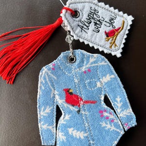 Puede incluir: Un adorno de tela con forma de camisa de manga larga con fondo azul claro, follaje de invierno blanco y un p&aacute;jaro cardenal rojo. Incluye una borla roja y una etiqueta blanca con el texto bordado Always with you y un cardenal.