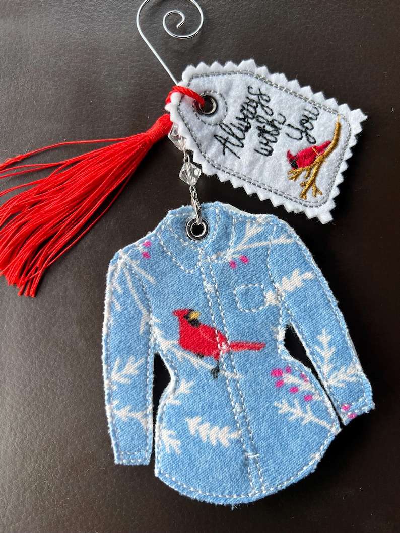 Puede incluir: Un adorno de tela con forma de camisa de manga larga con fondo azul claro, follaje de invierno blanco y un p&aacute;jaro cardenal rojo. Incluye una borla roja y una etiqueta blanca con el texto bordado Always with you y un cardenal.