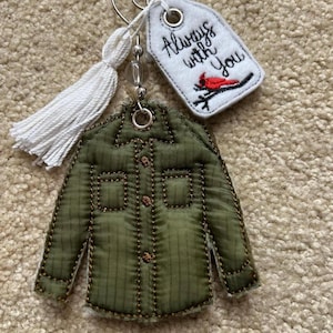 May include: A keychain featuring a quilted olive green shirt-shaped fabric piece, a white tassel, and a white fabric tag with the embroidered text Always with You and a red cardinal bird perched on a black branch, all attached to a metal ring.